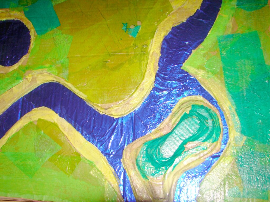 Obrazek pokazuje fragment makiety zbudowanej przez dzieci. Teren z tektury jest oklejony gładka bibuła w w kilku odcieniach zieleni. Rzeka ze staniolu intensywnie niebieska i połyskująca.