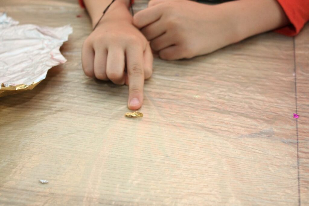 Dwie dziecięce dłonie oparte o blat stołu. Palec prawej dłoni wskazuje maleńki, nieregularny zwitek staniolu. To model samochodu dla budowanej makiety.