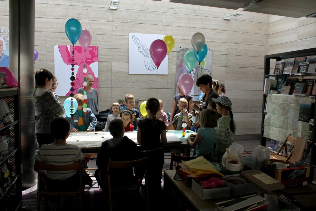 Grupa dzieci w rożnym wieku stoi wokół stołu. Na stole tektury do budowy makiety miasta
