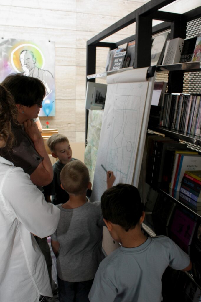 Prowadząca warsztaty i dzieci stoją przed rysunkiem planu miasta. Mały chłopiec z ołówkiem w dłoni coś tłumaczy.